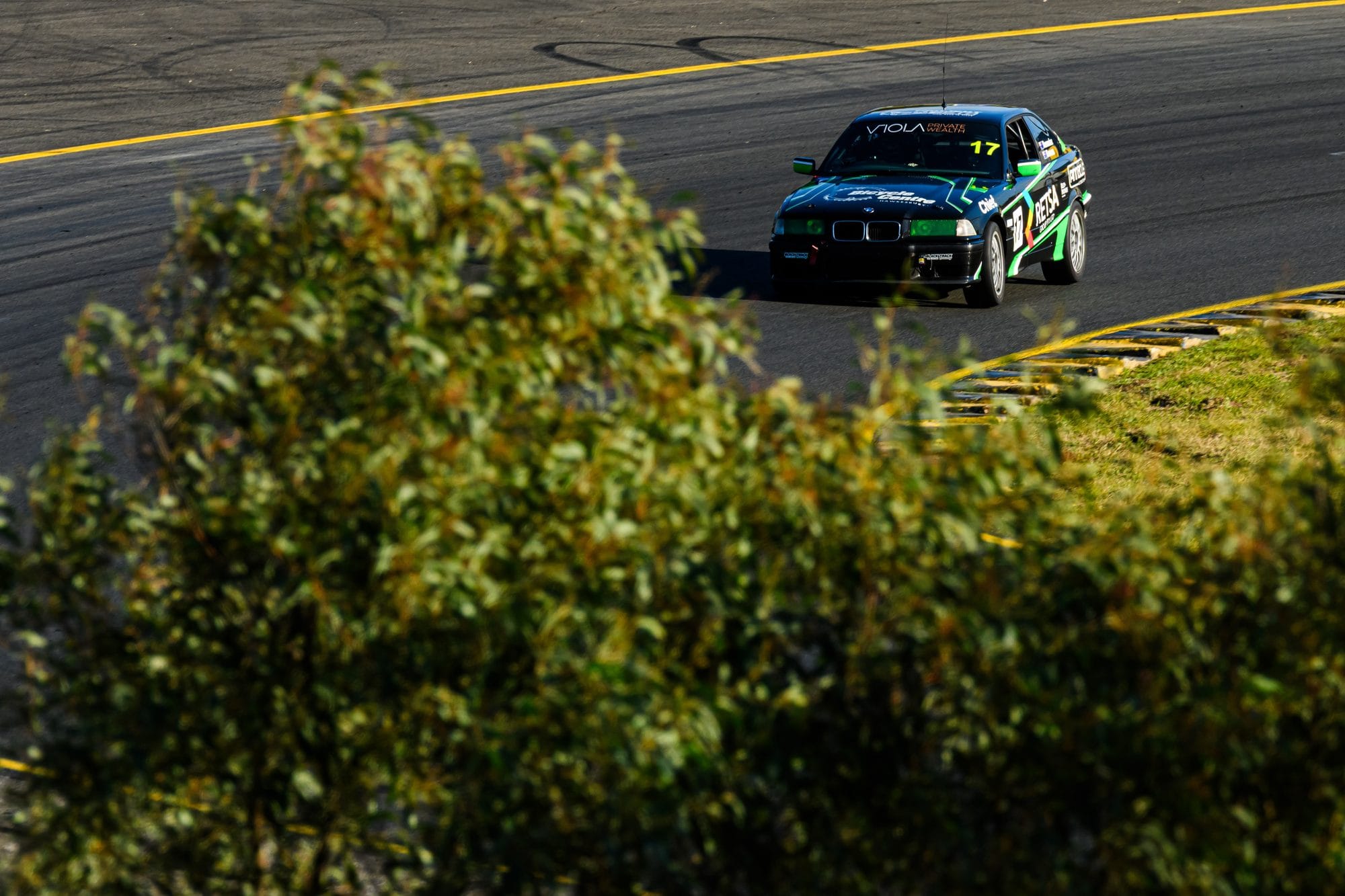 The #17 Project E36 BMW 328i cornering in warm sunlight, framed through trackside foliage at Sydney Motorsport Park during MRA Round 5